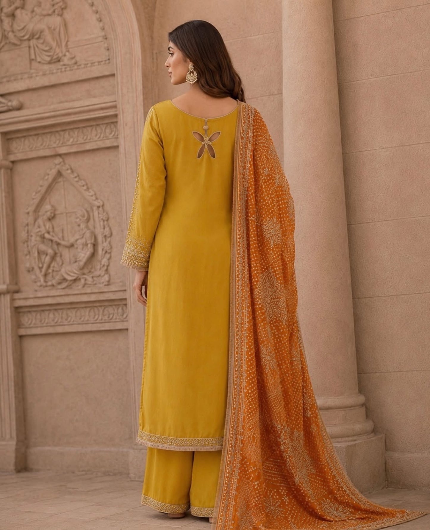 A woman wearing a mustard yellow traditional salwar kameez suit with matching trousers and a bright orange patterned dupatta, standing in front of a beige stone archway.