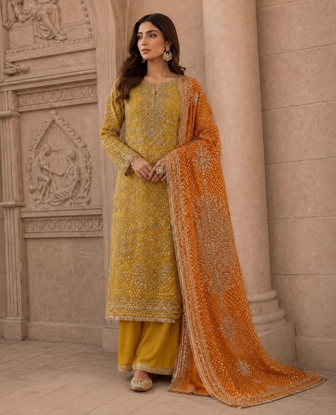 A woman models a traditional Indian salwar kameez suit set consisting of a mustard yellow embroidered kurta, matching yellow trousers, and a bright orange dupatta with intricate gold embroidery, set against a classical stone column background.