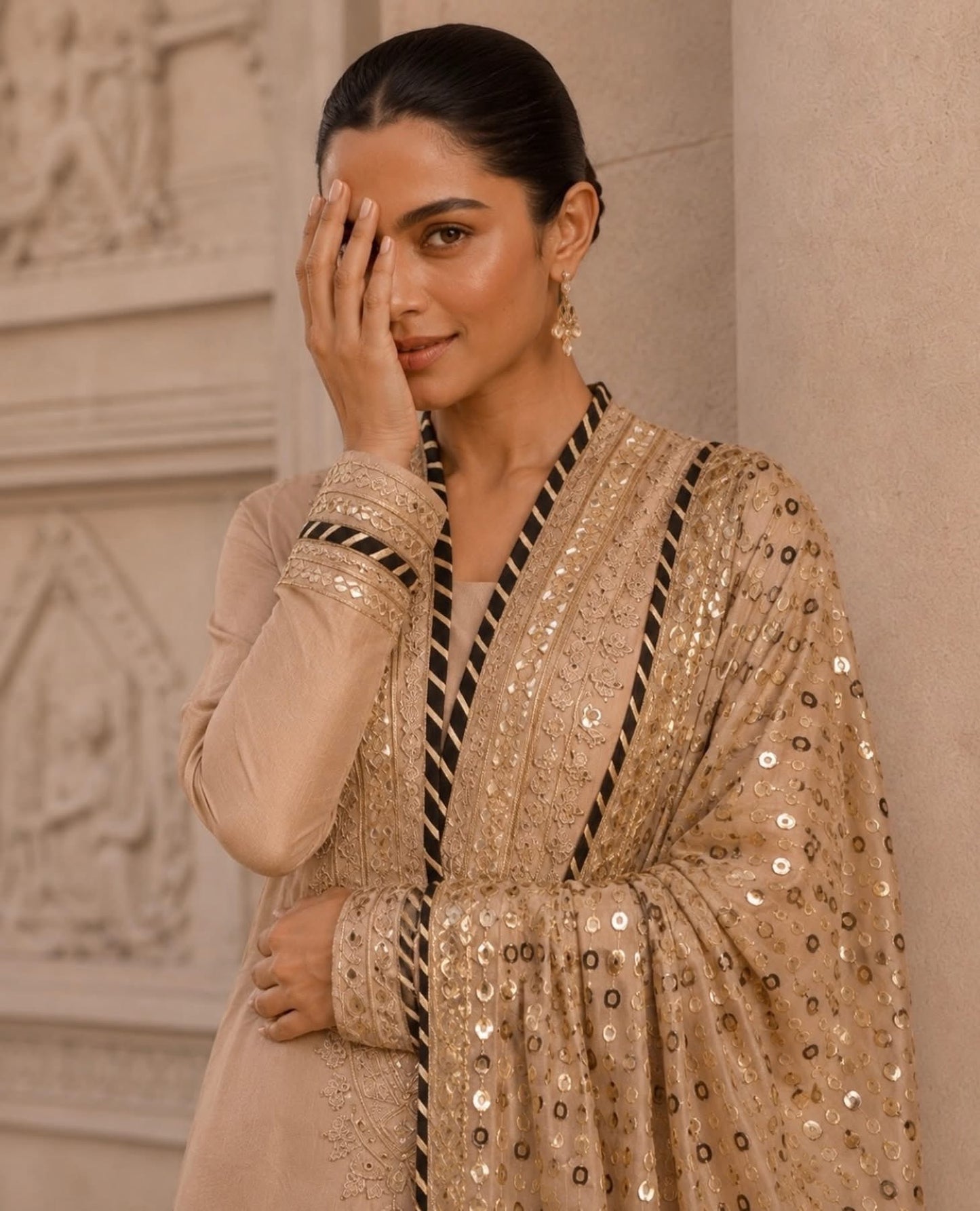 A woman wearing a beige traditional outfit with black and gold embroidered details and a matching sheer draped shawl, posed against a light stone wall.