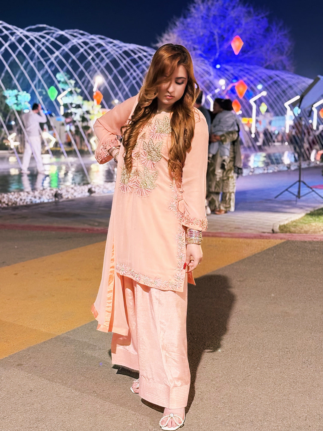 Woman in a peach traditional outfit standing in front of a decorated fountain at night.
