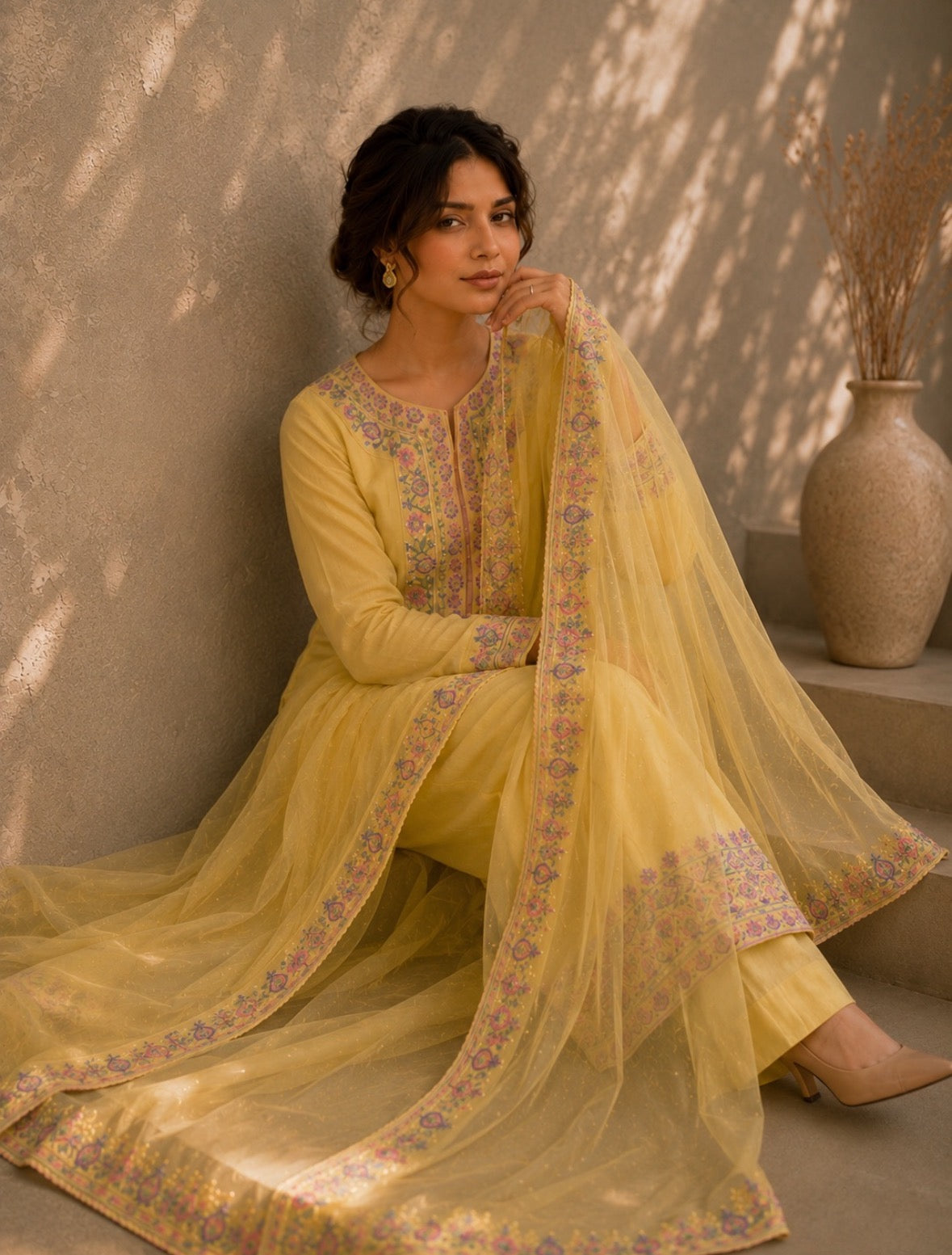Woman in a yellow traditional outfit with floral patterns sitting against a neutral wall.