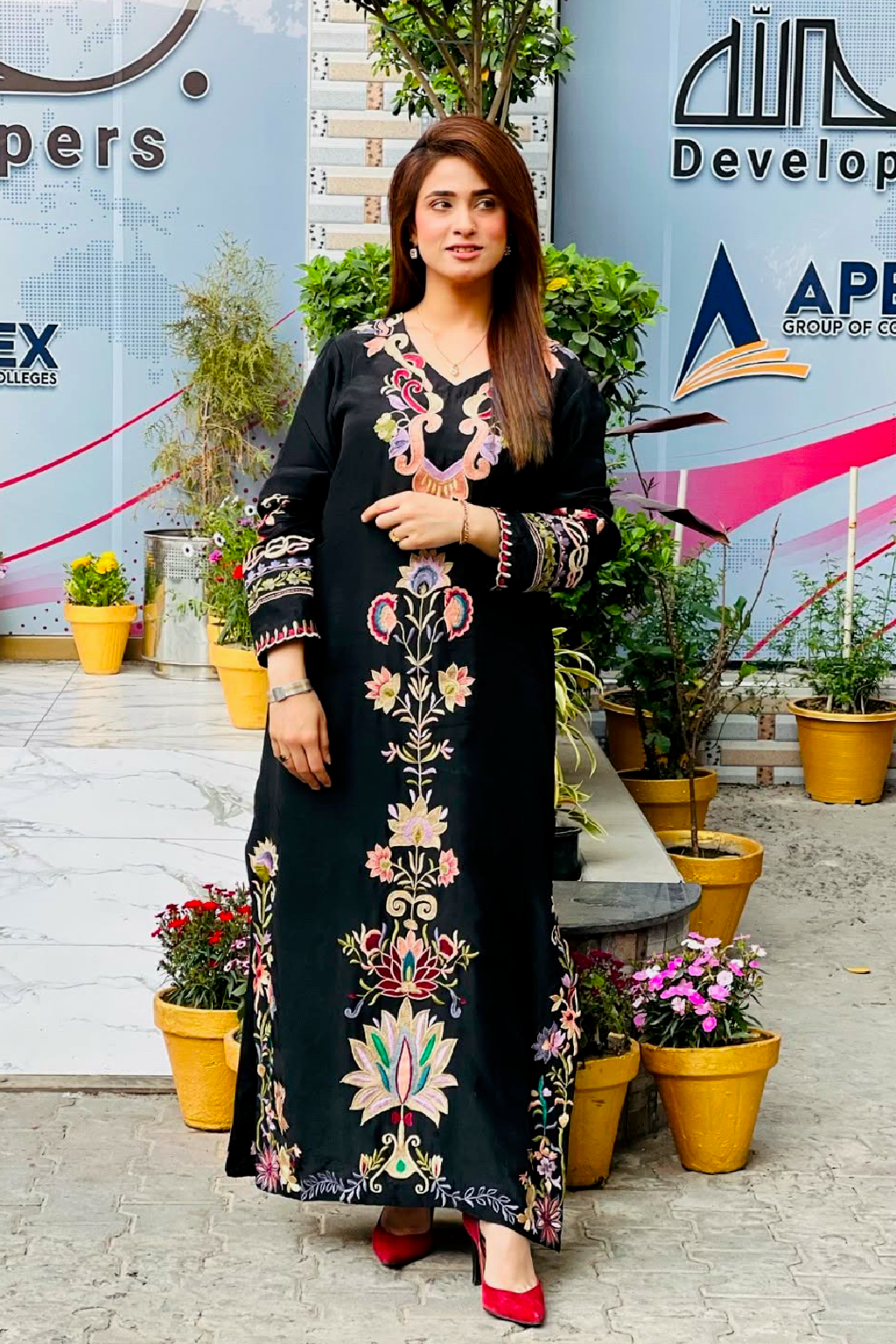 Woman in a black dress with floral embroidery standing outdoors with plants and a building in the background.