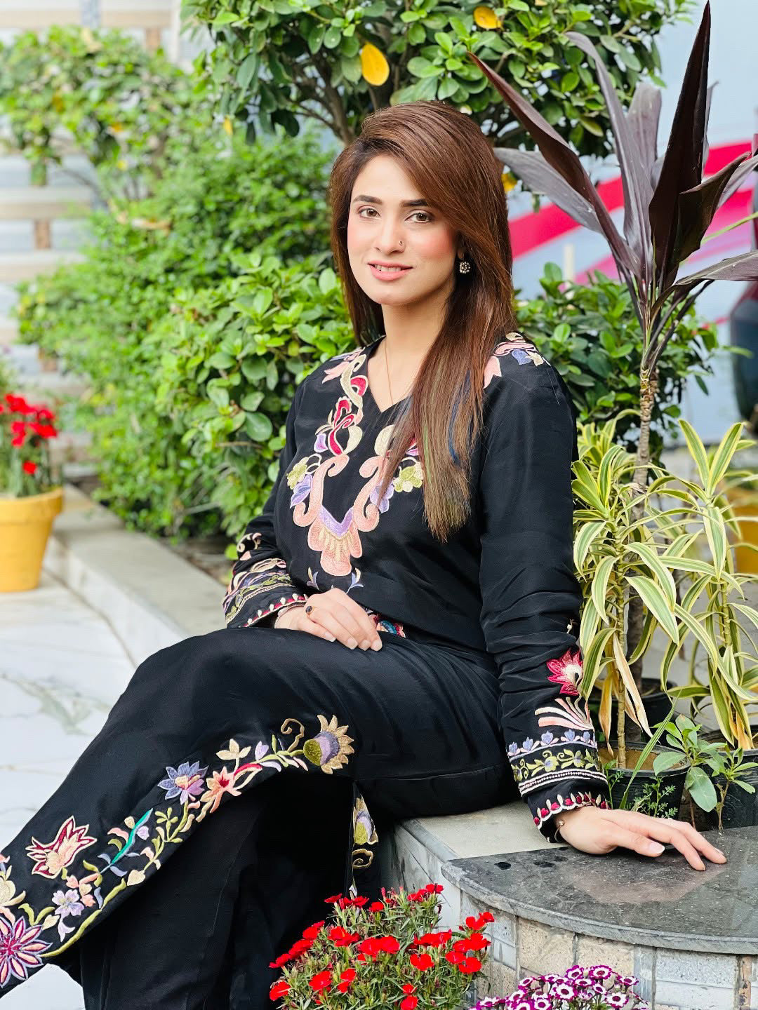Woman in black embroidered outfit sitting outdoors with plants and flowers around