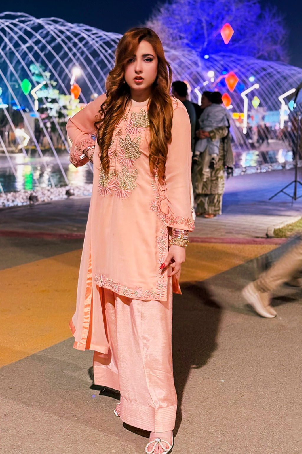 Woman in a peach embroidered outfit standing in front of a decorative fountain at night.
